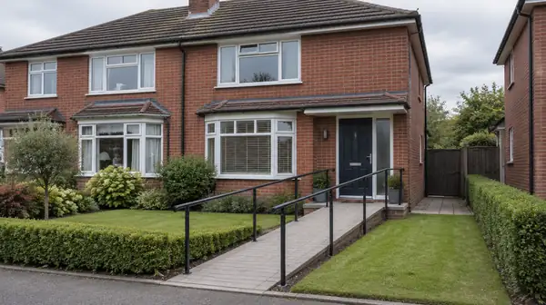 A paved ramp with black steel railings crossing the front garden of a red-brick semi-detached house to its front door.