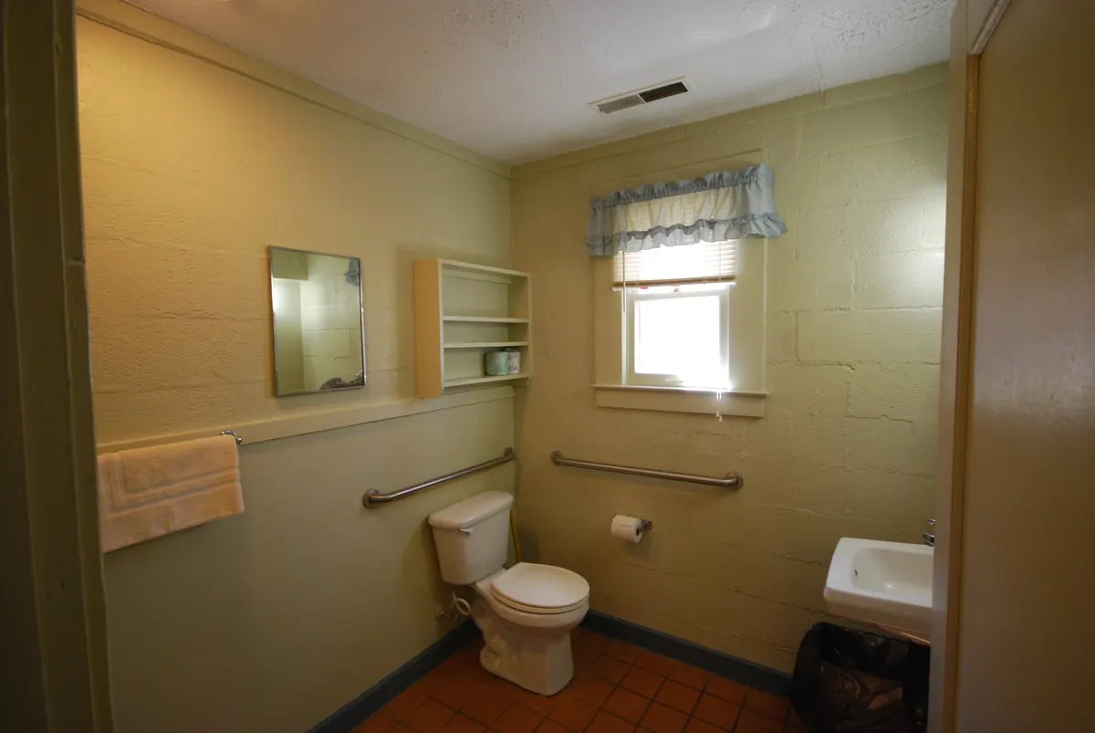 A small bathroom with grab bars on the side wall and the back wall of a toilet, a wall-hung sink and a window.