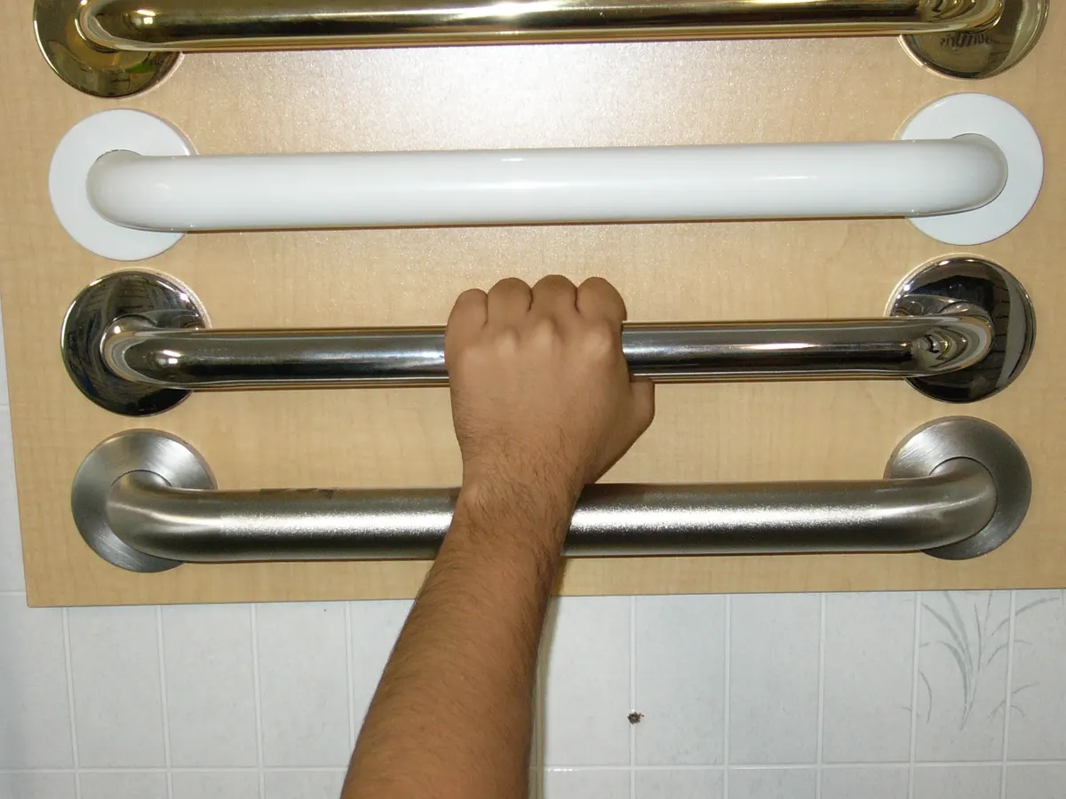 A hand gripping one of four grab bars of different finishes and diameters mounted on a display board.