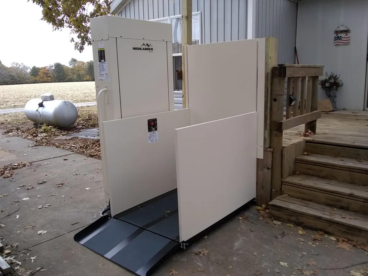 A vertical platform lift with a hinged gate and a short entry ramp beside the wooden steps and deck of a house.
