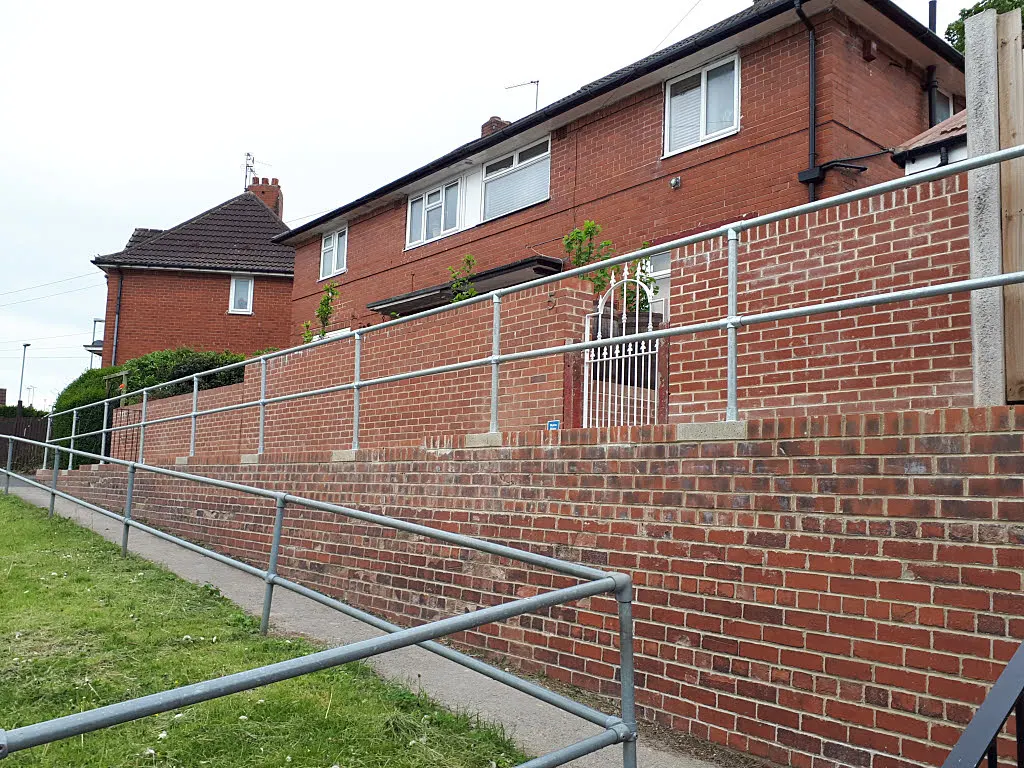 A long ramp with metal handrails running beside a brick retaining wall up to the front of a semi-detached house.