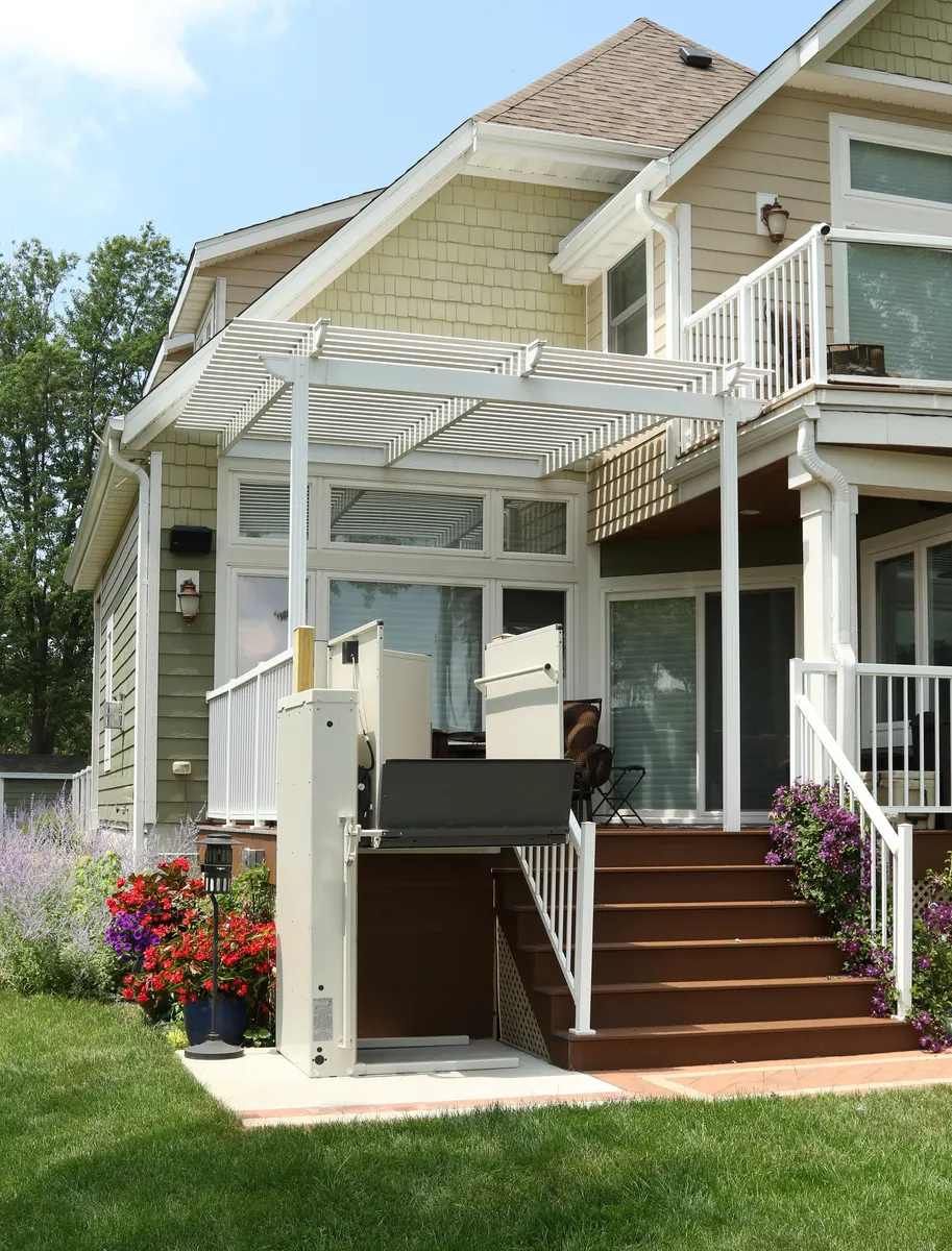 A vertical platform lift with a gate beside the front steps of a two-storey house with a covered porch.