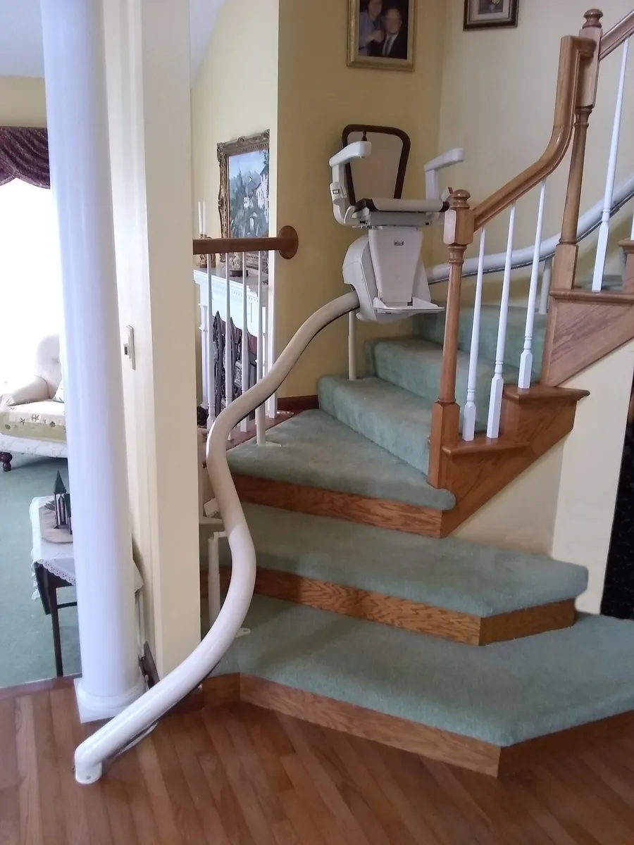 A stair lift chair with folded armrests on a curved rail that wraps around the bottom of a carpeted turning staircase in a house.