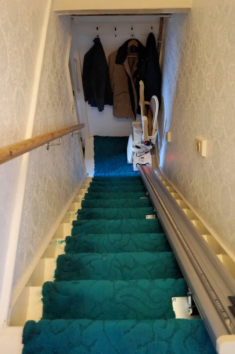 A stair lift rail running down one side of a narrow carpeted staircase in a terraced house, with the folded chair parked at the top.