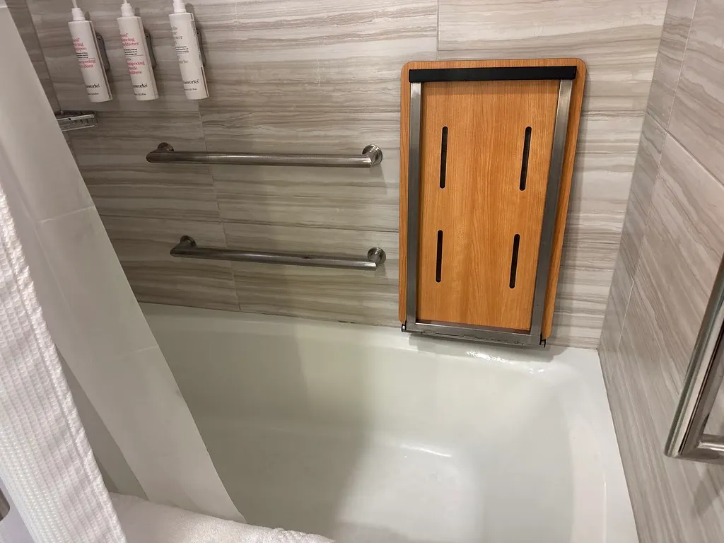 Two horizontal metal grab bars on the tiled back wall of a bathtub, one above the other, with a wooden fold-down seat on the end wall.